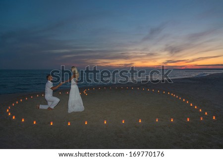A man propose marriage to woman in candles heart at sea beach against sunset- St.Valentines Day romantic concept