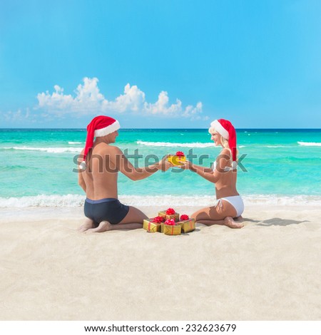 Happy couple at sea beach in santa hats give presents for each other - christmas or new year holidays in hot countries concept