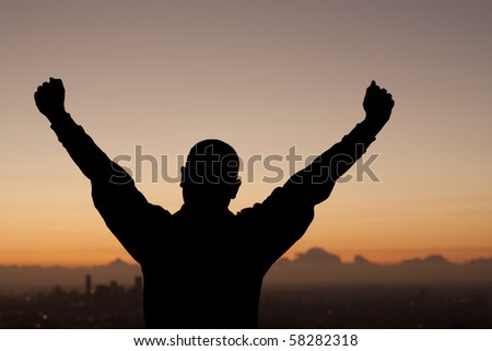 Silhouette of man above city at sunrise with victorious arms.
