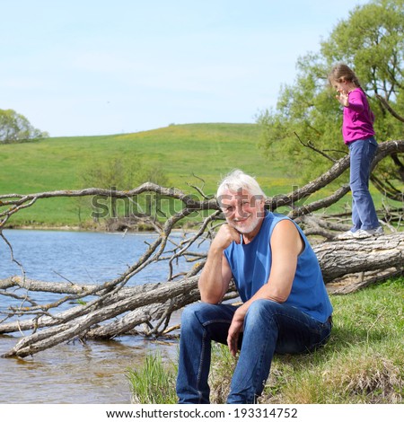 Pensive smiling grandfather and funny granddaughter near the fallen tree on the river bank.Grandfather and granddaughter travel to natural places