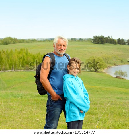 Portrait of grandfather with a travel backpack and his granddaughter on background a spacious landscape. Grandfather and granddaughter travel to natural places
