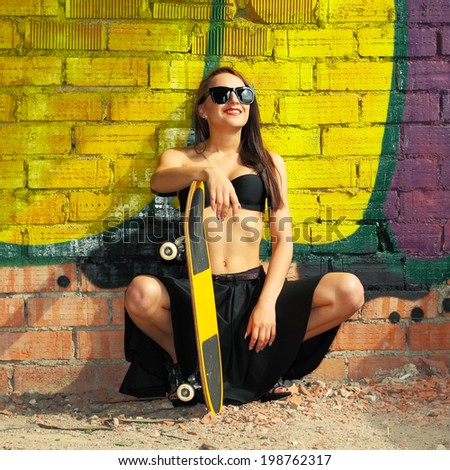 Sexy girl in sunglasses in top and skirt posing with a skateboard on the background wall of graffiti. Lifestyle outdoor portrait of girl in sunglasses