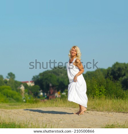 Mature beautiful blonde in a white dress walks on the sandy road on blue sky background