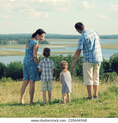 happy young family of four having fun on the top of a hill on a background of clouds. family of four standing on a hill and looking at spaces. View from the back.