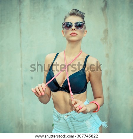 Stylish fashionable hipster blonde in sunglasses with a pink belt posing on wall background