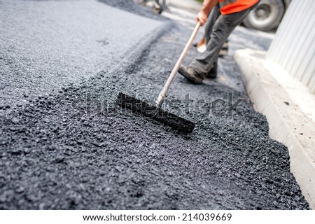 Worker leveling fresh asphalt on a road construction site, industrial buildings and teamwork