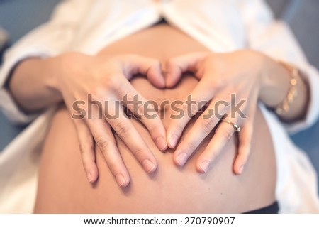 Pregnant young woman holding fingers in a heart shape, touching belly on baby bump and loving the baby. Mother love concept.