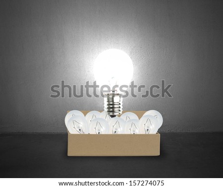 Big glowing light bulb float over opened cardboard box with many smaller bulbs in concrete wall background
