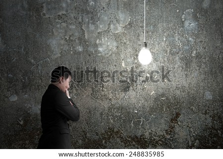 Businessman pondering with brightly light bulb illuminated dark concrete wall  background