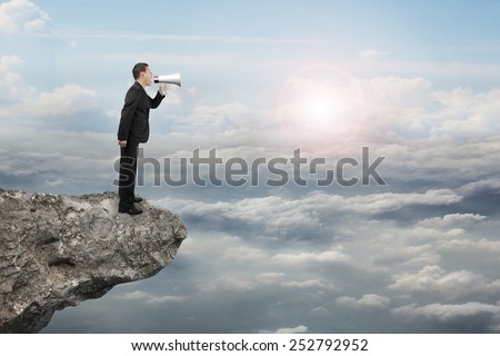 Businessman using megaphone yelling on cliff with sunlight cloudscape background
