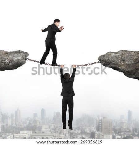Businessman walking on the cracking rusty iron chains another man holding connect two cliffs with urban scene skyline background, business teamwork concept.