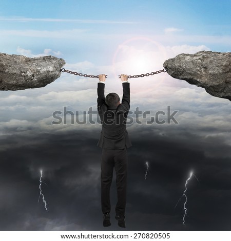 Businessman hand hanging on the cracking rusty iron chains connect two cliffs with opposite weather conditions sky background, bright sun cloudscape and dark cloudy lightning.