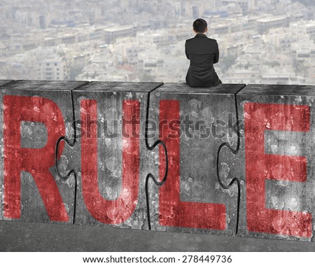 Business man sitting on four huge concrete puzzles with red rule word, on city landscape background