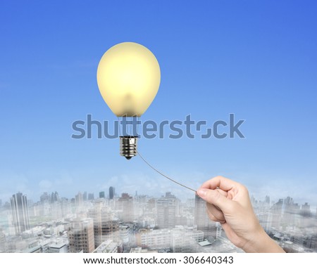 Woman hand pulling rope connected yellow lightbulb shape hot air balloon, with sky urban scene background.