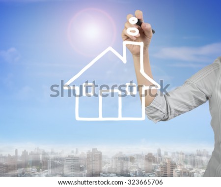 Female hand holding pen drawing house shape clouds in sky, front view, with sunny cityscape background.
