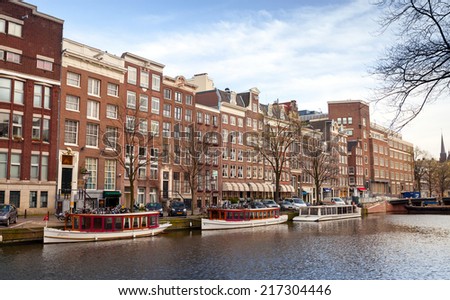 AMSTERDAM, NETHERLANDS - MARCH 19, 2014: Colorful houses and pleasure boats stand moored along canal embankment. Ordinary people walk on the coast