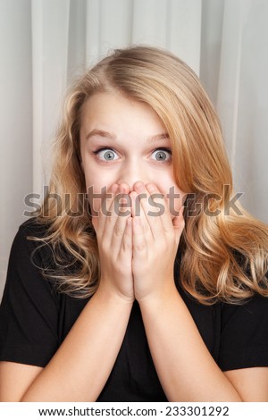 Beautiful blond Caucasian surprised girl opened her eyes wide and covers her mouth with her hands, vertical closeup studio portrait