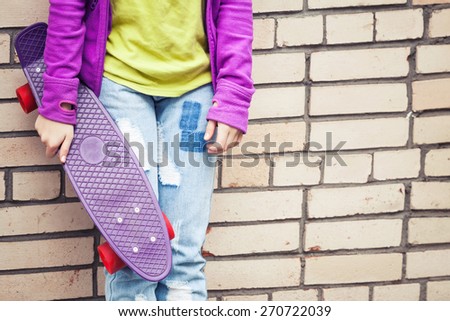 Blond teenage girl in jeans and colorful sporty clothes holds skateboard near by gray urban brick wall