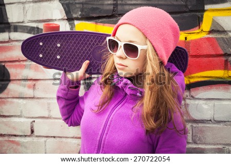 Blond teenage girl holds skateboard near by urban wall with graffiti