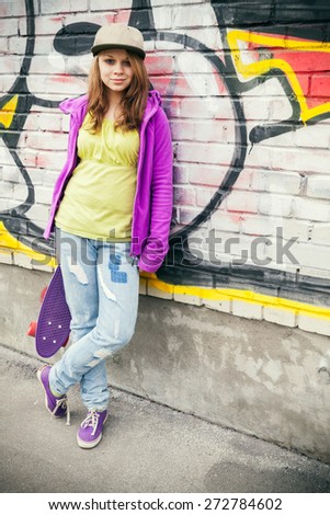 Blond beautiful teenage girl in cap holds skateboard near urban wall with colorful graffiti