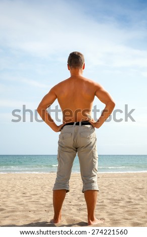 Young sporty sunburned barefoot man dressed in shorts stands on the sandy beach and staring at the sea, back view