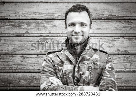 Young smiling Caucasian man in camouflage, outdoor black and white portrait over rural wooden wall