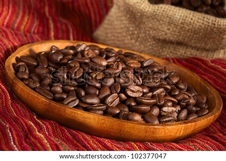Roasted coffee beans on wooden plate on red cloth with jute bag in the back (Selective Focus, Focus one third into the coffee beans)
