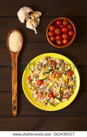 Overhead shot of rice dish with mincemeat and vegetables (sweet corn, cherry tomato, zucchini, onion) served on plate with ingredients around, photographed on dark wood with natural light