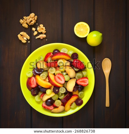 Overhead shot of fresh fruit salad made of grape, strawberry, plum and nectarine served on plate with walnuts, lemon and spoon beside, photographed on dark wood with natural light