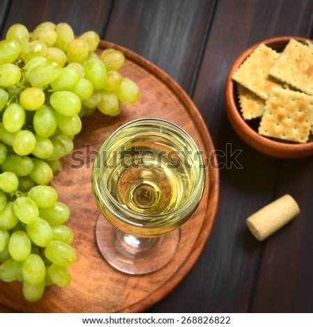 Glass of white wine with white grapes, crackers and a cork, photographed on dark wood with natural light (Selective Focus, Focus on the rim of the wine glass)