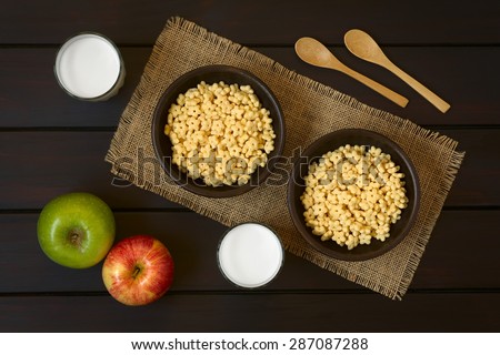 Honey flavored breakfast cereal in two rustic bowls with glasses of milk, apples and wooden spoons on the side, photographed overhead on dark wood with natural light