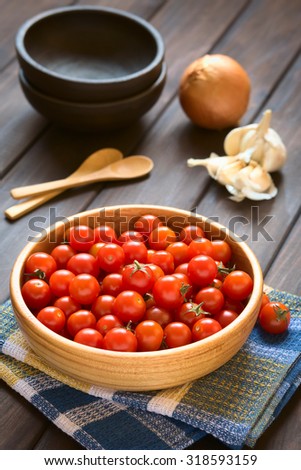 Cherry tomatoes in wooden bowl with garlic, onion, bowls and wooden spoons in the back, photographed on wood with natural light (Selective Focus, Focus one third into the tomatoes)