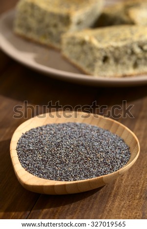 Raw poppy seeds on small wooden plate, poppy seed cake in the back, photographed with natural light (Very Shallow Depth of Field, Focus one third into the poppy seeds)