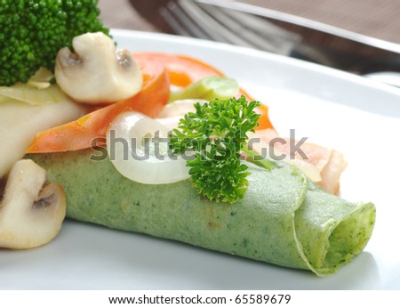 Rolled thin green colored pancake (colored with spinach in dough) with vegetables on top with fork in the background (Selective Focus, Focus on the front side of the pancake and the parsley)