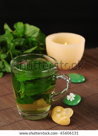 Fresh mint tea with cookies, green stones, a lit candle and mint leaves on dark brown table mat with black background (Selective Focus, Focus on the front of the glass, the handle and the cookie)