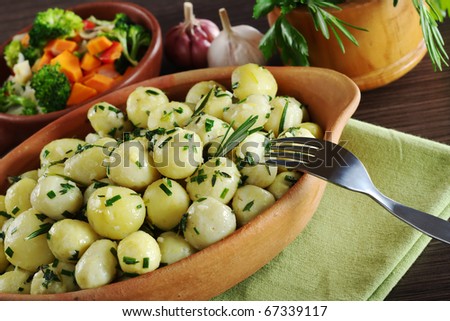 Small potatoes with herbs, such as parsley, thyme and rosemary with a fork; with fried vegetables, garlic and fresh herbs in a wooden mortar in the background (Selective Focus, Focus on the rosemary)