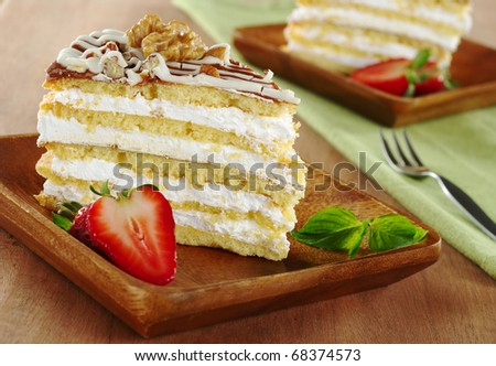 Nut cake garnished with a half walnut, a strawberry and a mint leaf served on a wooden plate with another cake and fork in the background (Selective Focus, Focus on the front of the cake and the nut)