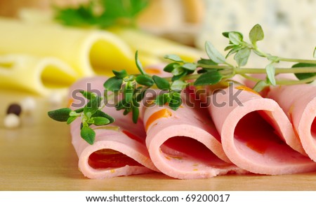 Cold cut slices on wooden board with thyme on top and cheese in the background (Selective Focus, Focus on the front of the cold cut slices and the tip of the thyme)