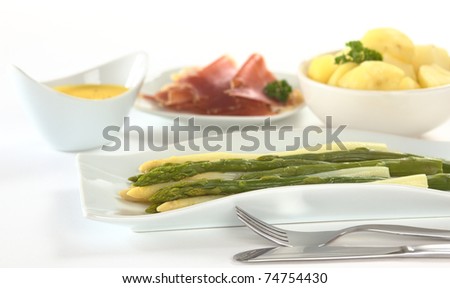 Cooked white and green asparagus on long plate with fork and knife in front and potatoes, ham and Hollandaise sauce in the background (Selective Focus, Focus on the front of the asparagus)