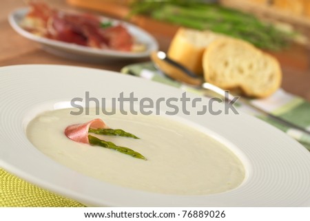 Cream of asparagus garnished with green asparagus heads and a slice of ham on top in a white soup plate (Selective Focus, Focus on the asparagus head in front and part of the ham slice on the soup)