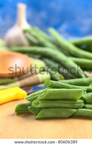 Raw cut green bush beans on wooden board with a kitchen knife, bean pods and garlic in the back (Selective Focus, Focus on the three beans lying on each other)