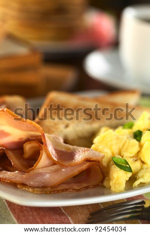 Fried bacon and scrambled eggs with toast bread, pancakes and coffee in the back (Selective Focus, Focus one third into the bacon and on the front of the eggs)