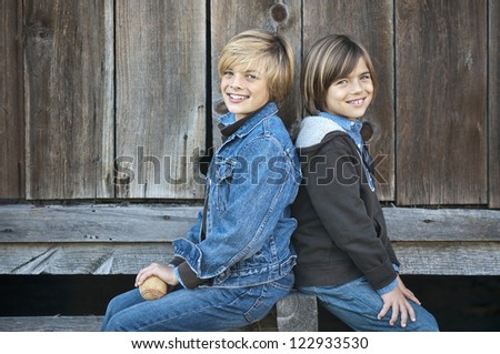 Two cute boys, brothers, sitting in front of an old barn, one holding a worn out baseball