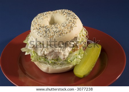 sesame seed bagel sandwich, chicken salad with bean sprouts,tomato,lettuce,cucumber, and a pickle, red plate on blue background
