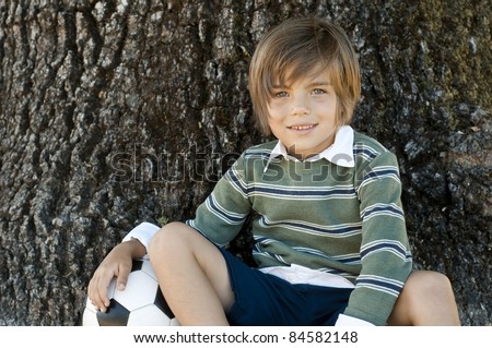 cute boy sitting by big oak tree, resting arm on soccer ball
