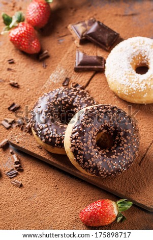 Chocolate and sugar donuts with fresh strawberries and dark chocolate served on cutting board with cocoa powder as background. See series