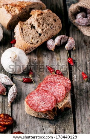 Sandwich with sliced salami served with other sausages, fresh bread, garlic and red hot chili peppers on old wooden table. See series