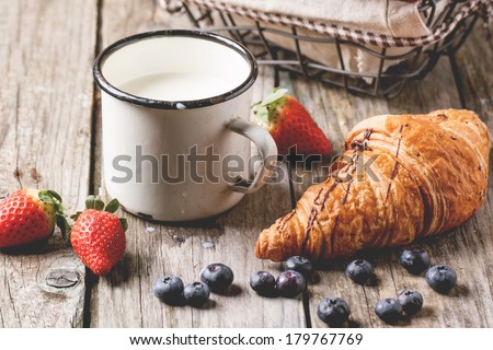 Fresh croissant with vintage mug of milk and berries over old wooden table. See series.