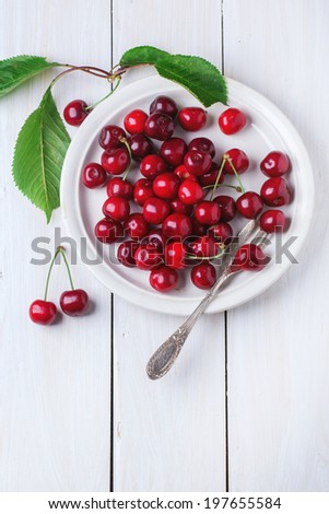 Ceramic plate of fresh cherries with leaves, served with dessert fork over white wooden table. Top view.