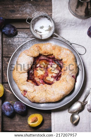Cake galette with plums, served in vintage metal plate over old wooden table. Top view. See series